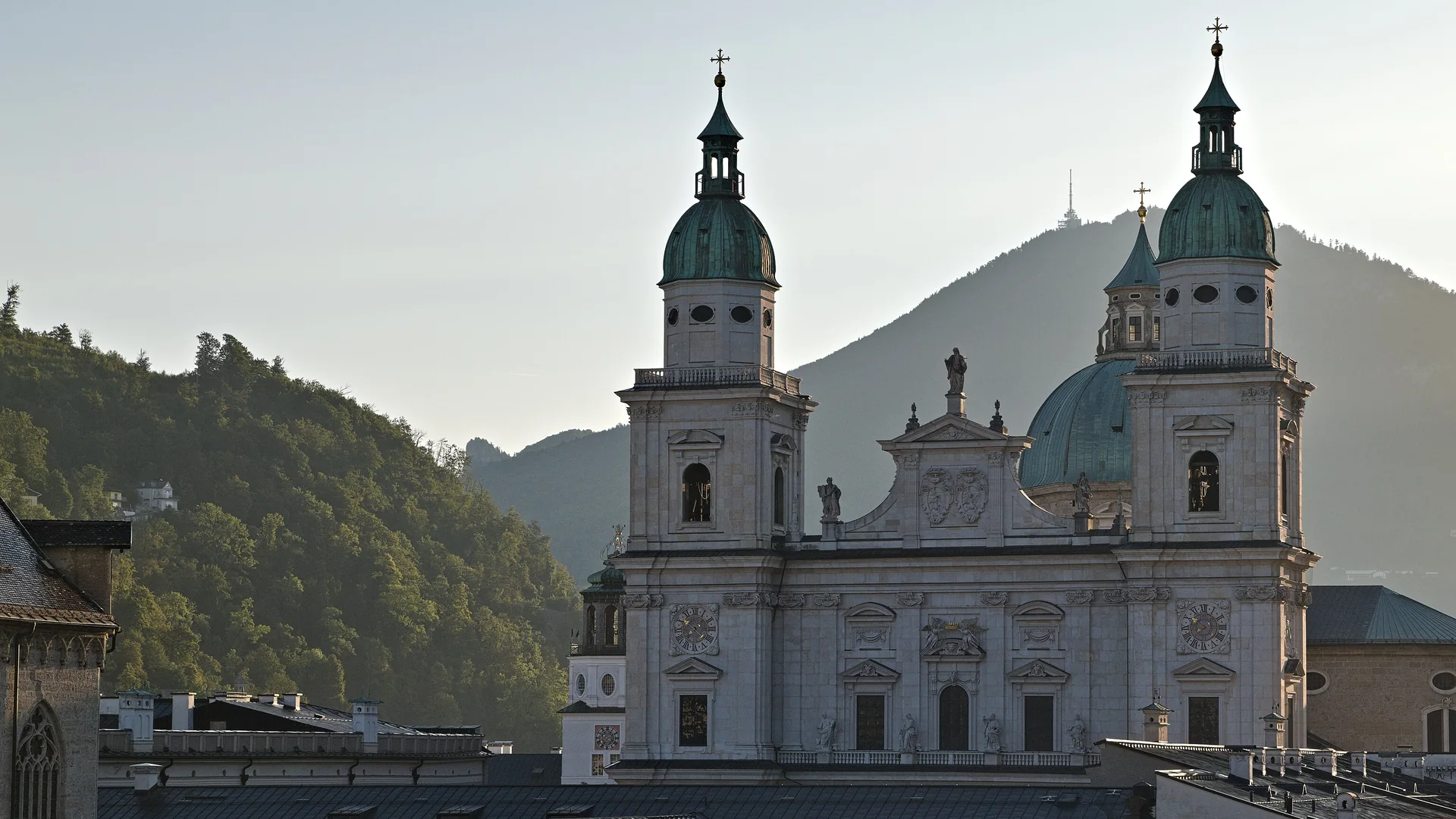 Die beiden Türme mit grünen Kupferhauben und die helle Barockfassade des Salzburger Doms über den Dächern der Altstadt, dahinter ein bewaldeter Berg im Morgenlicht