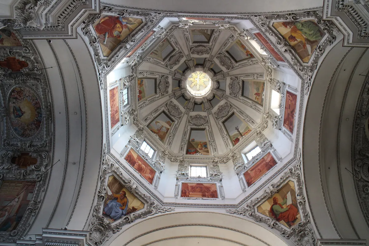 Der Blick hinauf in die achteckige Kuppel des Salzburger Doms mit bemalten Feldern, Stuckarbeiten und einer goldenen Taube im Zentrum