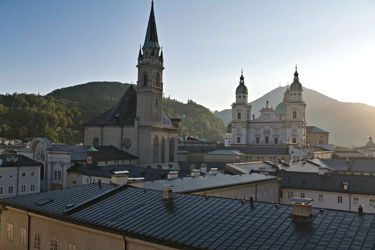 Der Dom und die Franziskanerkirche gemeinsam über den Dächern der Salzburger Altstadt, mit einem bewaldeten Hügel im Hintergrund