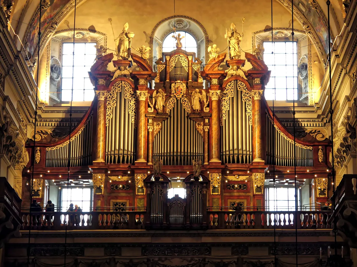 Die große Orgel auf der Westempore des Salzburger Doms, ein hohes Gehäuse mit vergoldeter Schnitzerei, geschnitzten Figuren obenauf und silbernen Pfeifen in warmem Licht