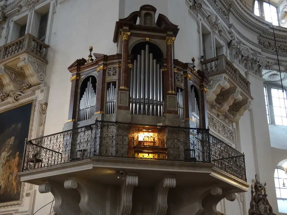 Ein Orgelgehäuse auf einer Empore an einem Pfeiler des Salzburger Doms, mit vergoldeten Kapitellen, silbernen Pfeifen und einem eisernen Geländer vor der Klaviatur