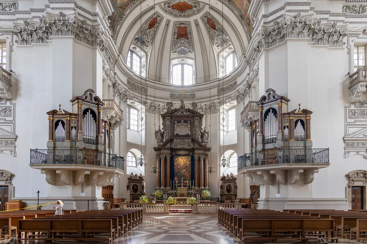 Das Langhaus des Salzburger Doms mit Blick zum Altar, mit je einer Emporenorgel auf den beiden vorderen Pfeilern unter der Kuppel und darunter Reihen hölzerner Kirchenbänke