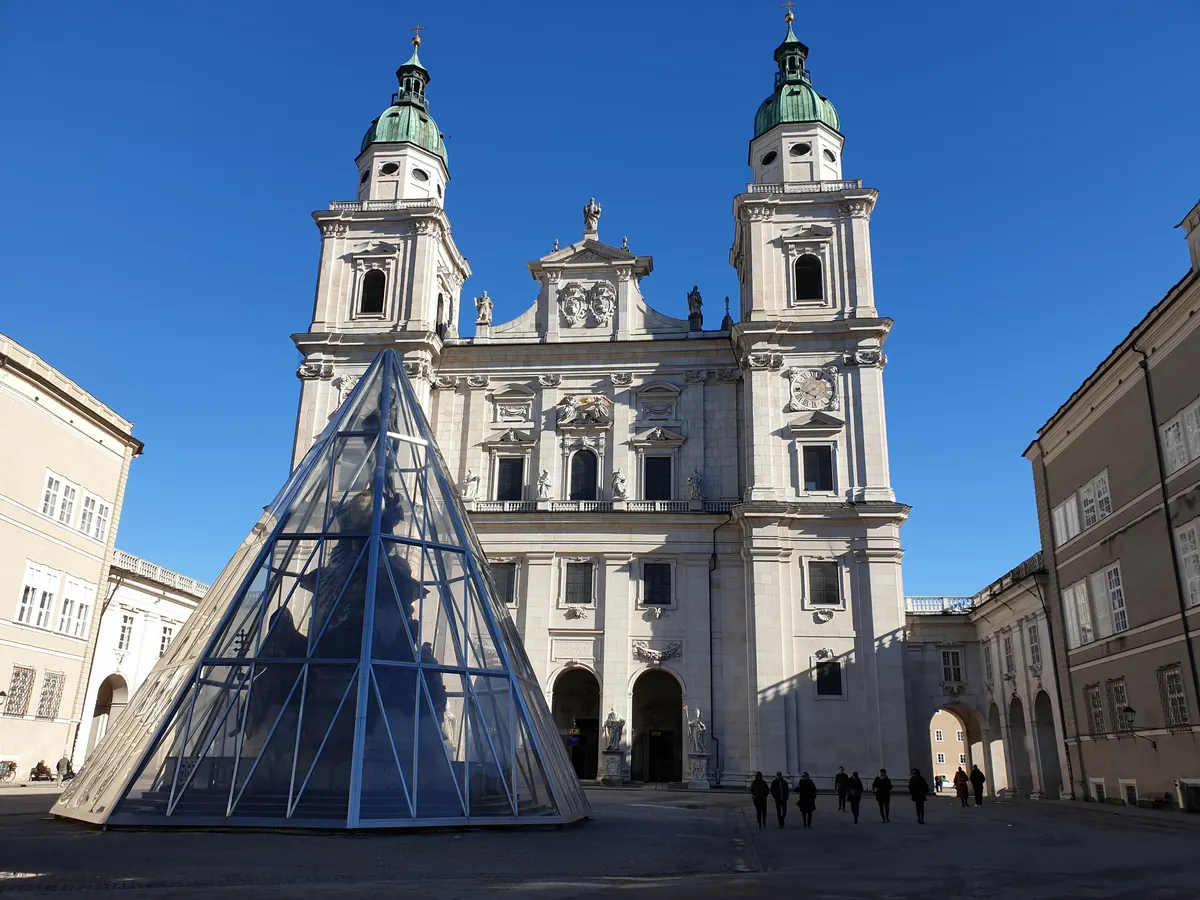 Die Westfassade des Salzburger Doms am Domplatz unter blauem Himmel, mit einer Glaspyramide über der Säule auf dem Platz und den Arkaden rechts